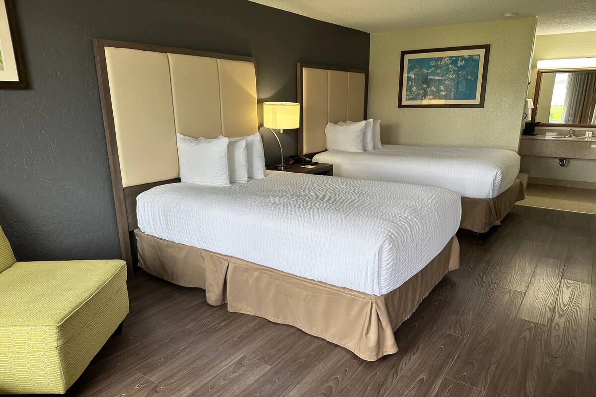 A hotel room with two beds, a bedside table with a lamp, a chair, a bathroom with a sink, and a painting on the wall.
