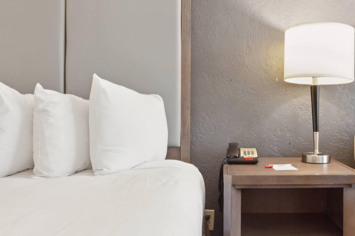 A hotel room with a bed, pillows, a bedside table with a lamp, a phone, and a notepad.