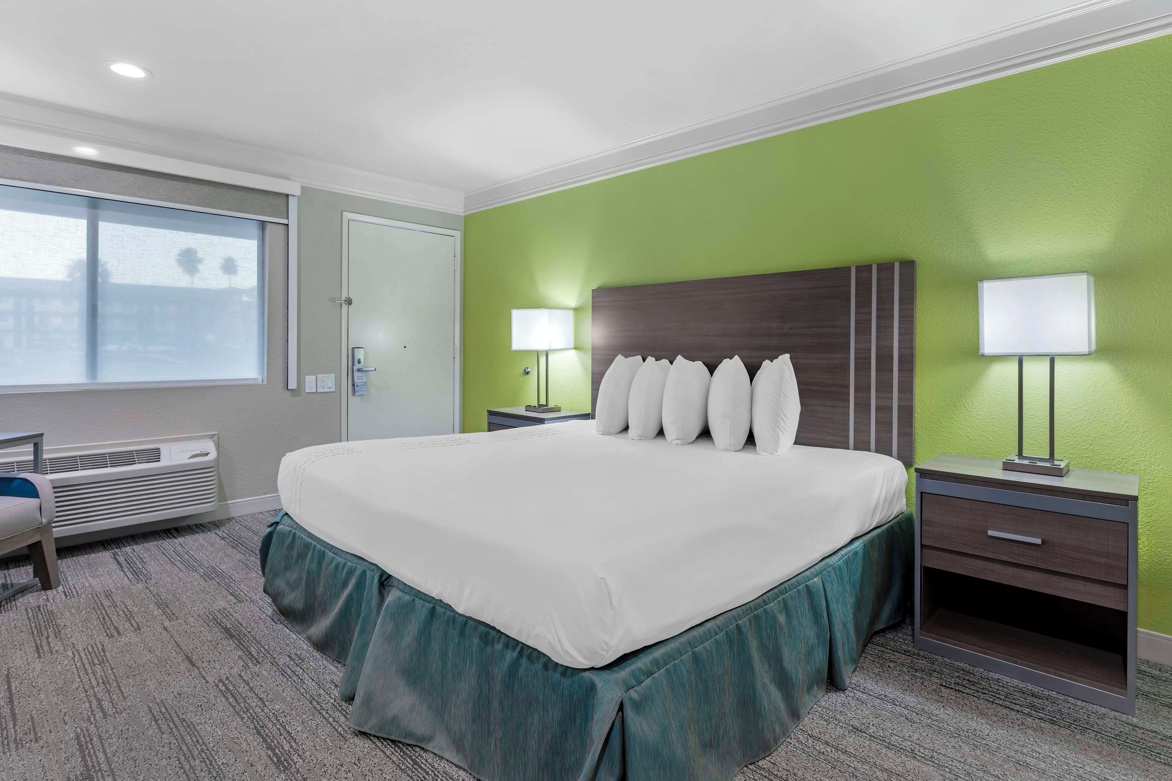 Quarto de hotel com uma cama grande, uma escrivaninha e uma cadeira. Há uma janela com persianas, uma mesa de cabeceira com abajur e um aquecedor fixado na parede.