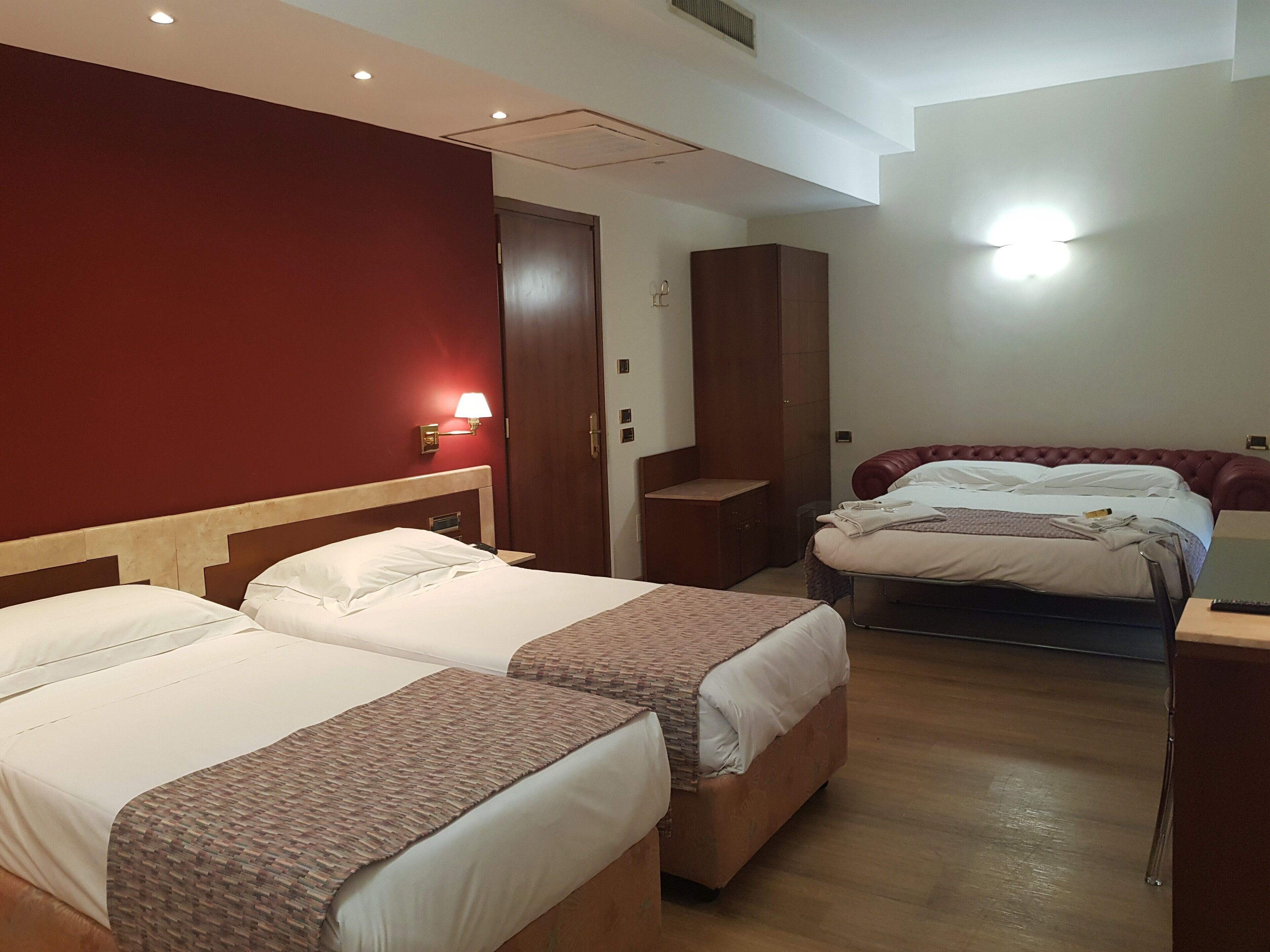A hotel room with two beds, a wooden headboard, a red accent wall, a nightstand, and a lamp.