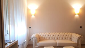 A living room with a beige sofa, two ottomans, a glass table, and two wall-mounted lights. - Hotel Astra Ferrara (Ferrara)
