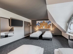 Una habitación de hotel moderna con dos camas individuales, un espejo grande, un televisor de pantalla plana y un cuadro de un edificio histórico.