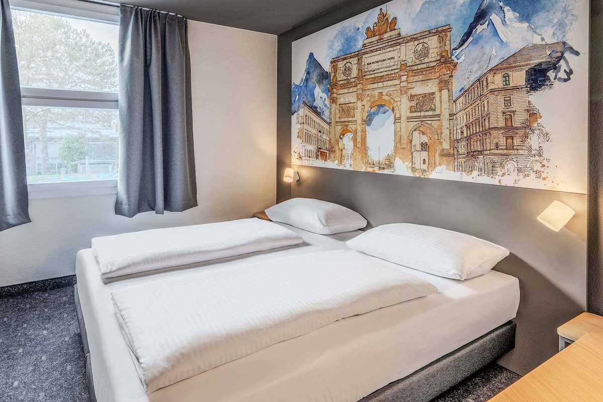 Una habitación de hotel con una cama, una ventana con cortinas, una lámpara de pared y un mural grande de un edificio histórico.