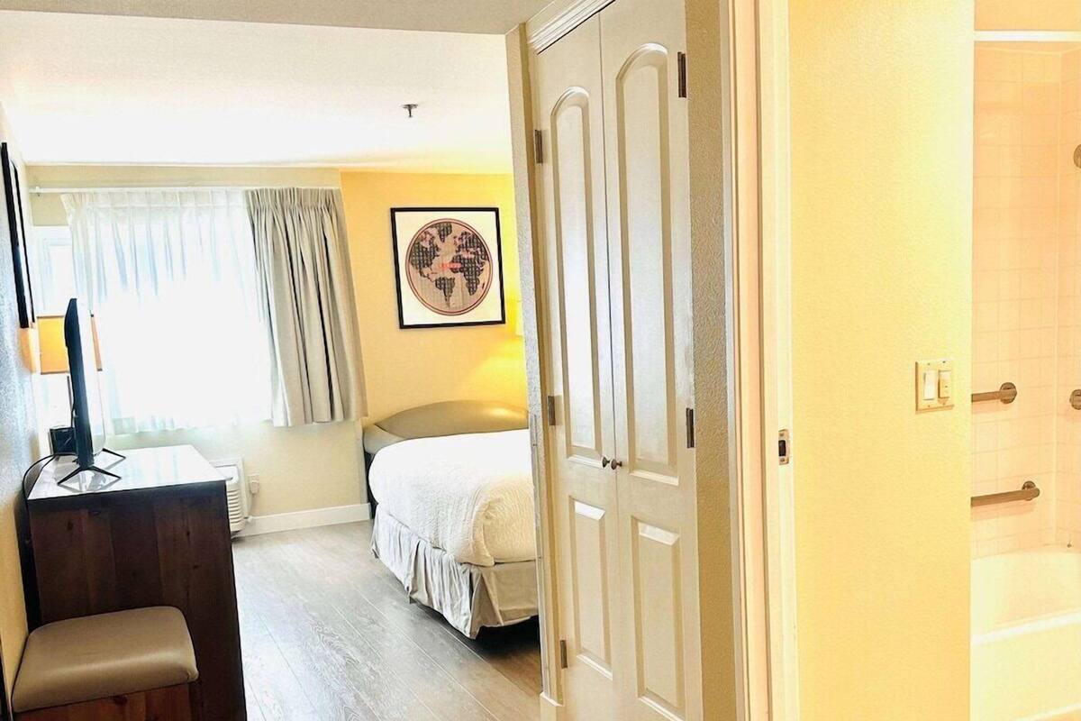 A hotel room with a bed, a desk with a computer, a chair, a window with curtains, and a door.