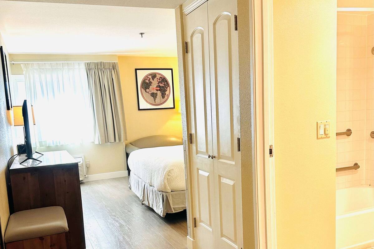 A hotel room with a bed, a desk with a computer, a chair, a window with curtains, a framed picture on the wall, and a door leading to a bathroom with a bathtub.