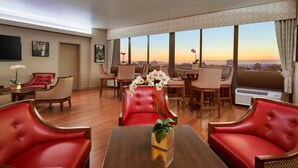 Ein Hotelzimmer mit Essbereich, roten Ledersesseln, einem Holztisch und einem großen Fenster mit Blick auf die Stadt.