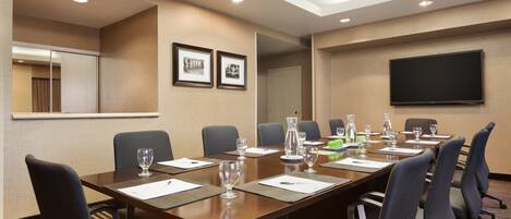 A conference room with a long table, multiple chairs, a flat-screen TV, framed pictures on the wall, and a mirror.