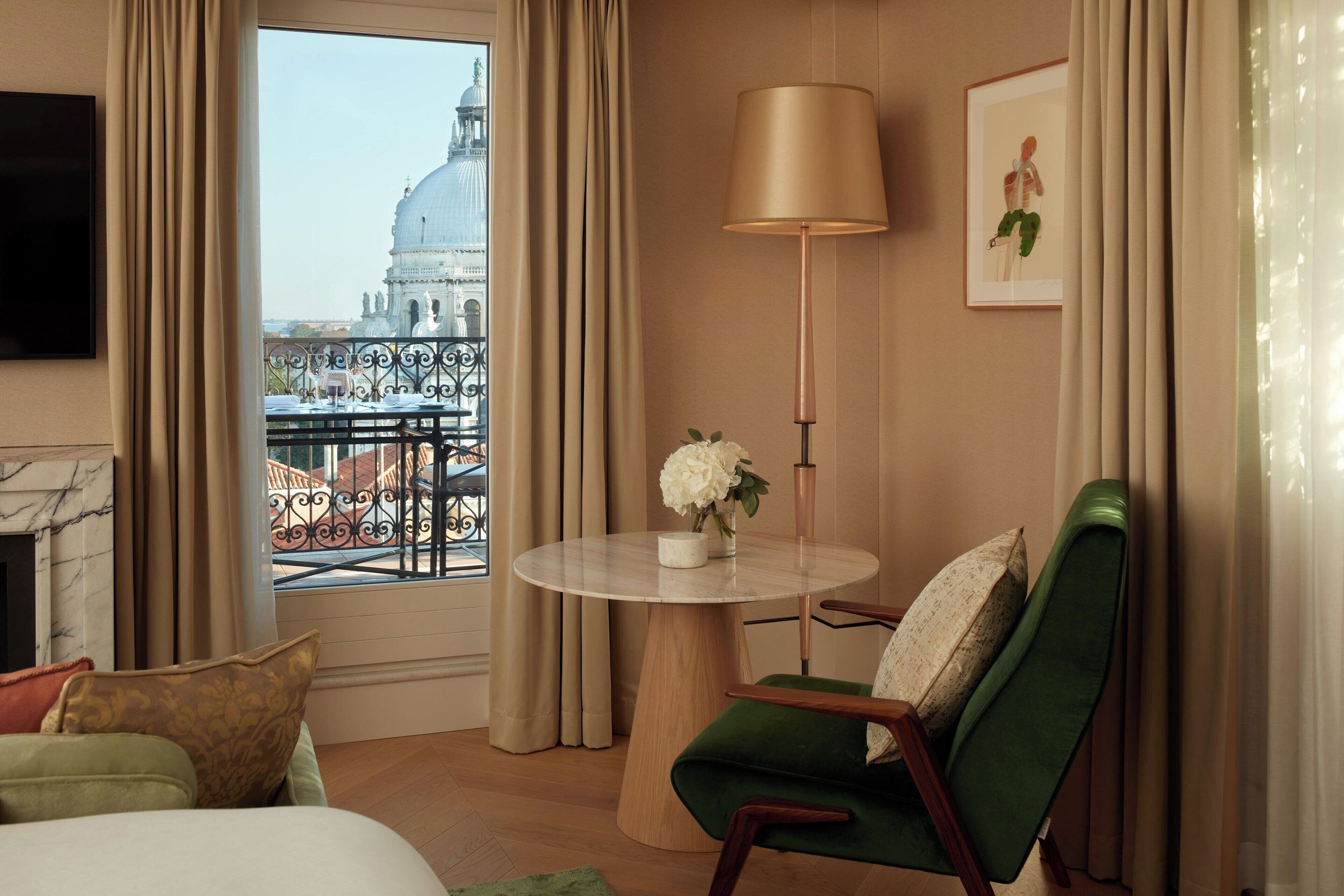 Een hotelkamer met uitzicht op een historisch gebouw, een ronde tafel met een bloevas, een groene fauteuil en een vloerlamp.