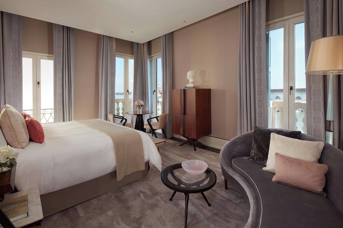 A hotel room with a large bed, a sofa, a small table, and a cabinet. There is a balcony with a railing and a view of the city.