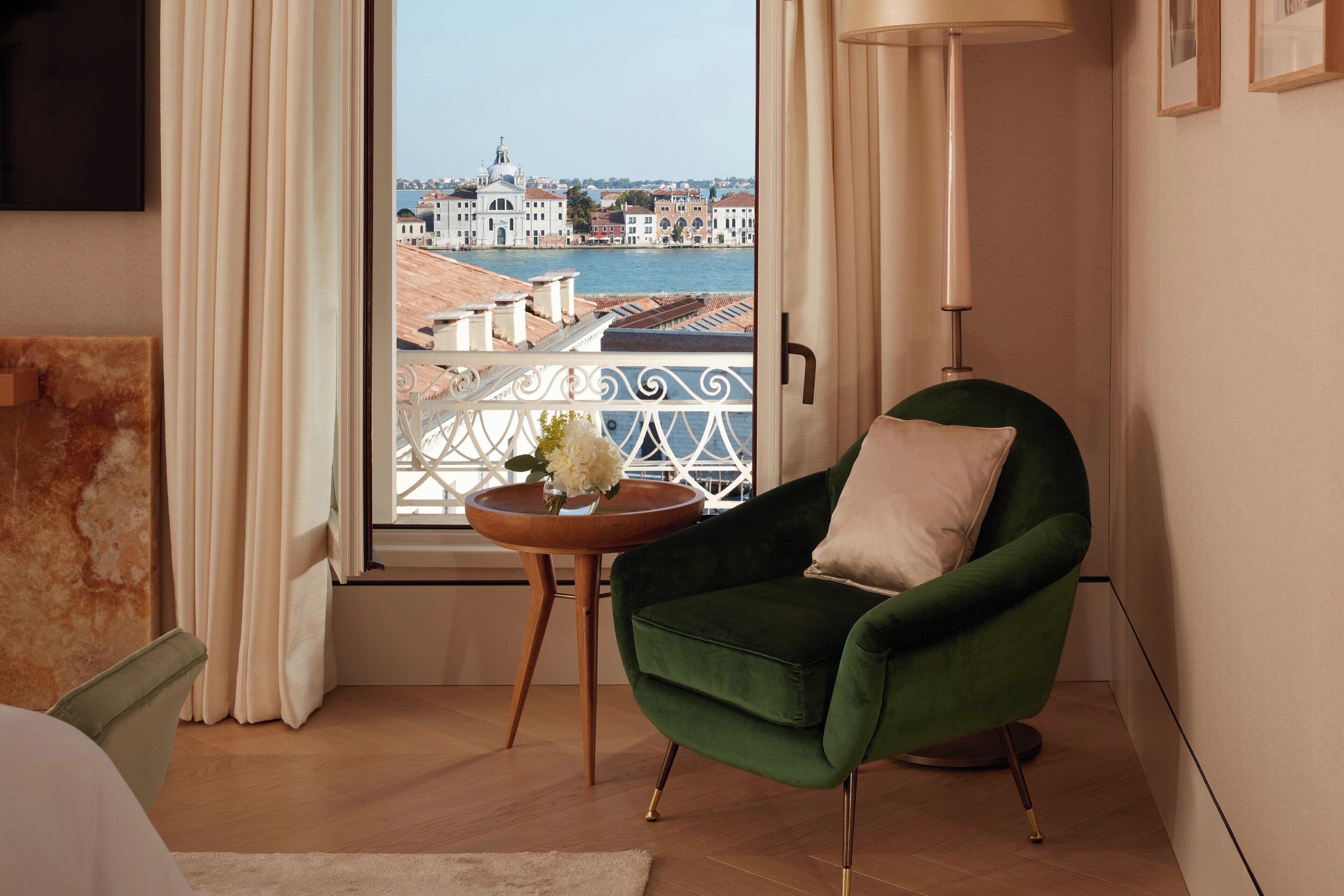 A room with a green armchair, a small wooden side table with a vase, and a view of a cityscape through a window with white curtains.