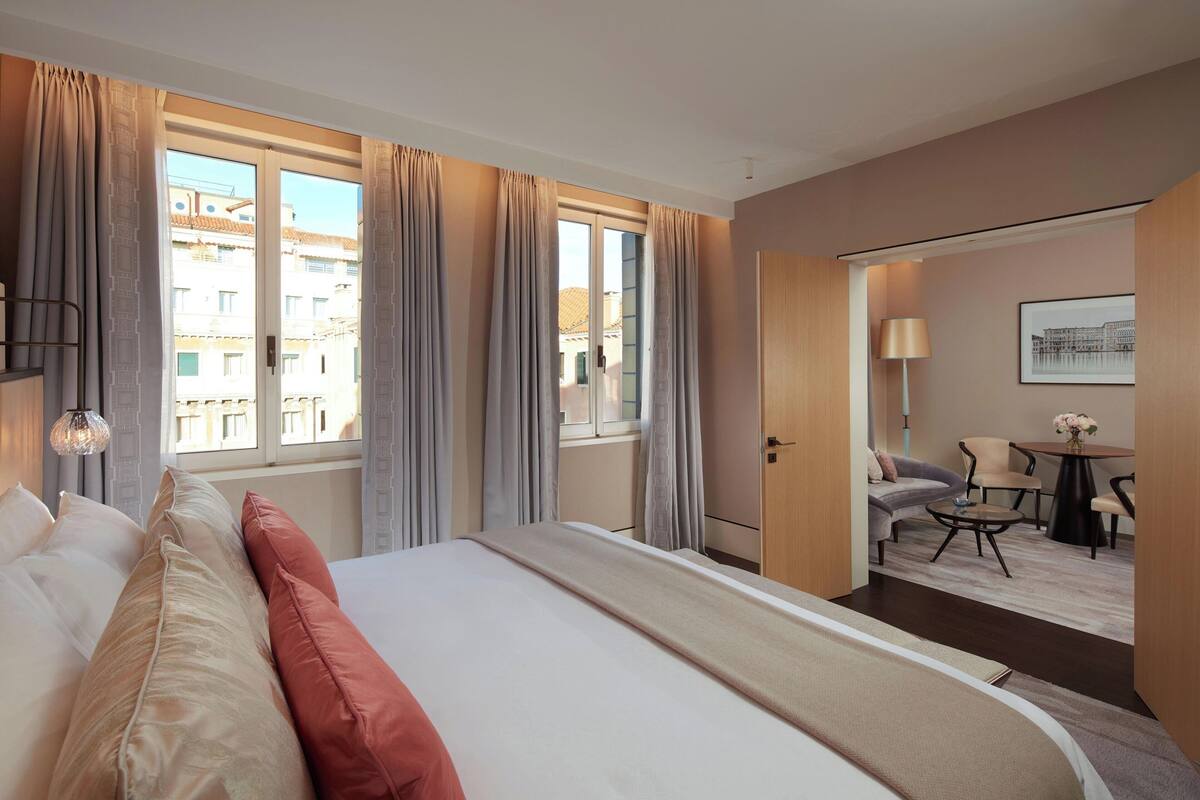 A hotel room with a large bed, a desk, and a chair. There is a window with curtains, a lamp, and a framed picture on the wall.