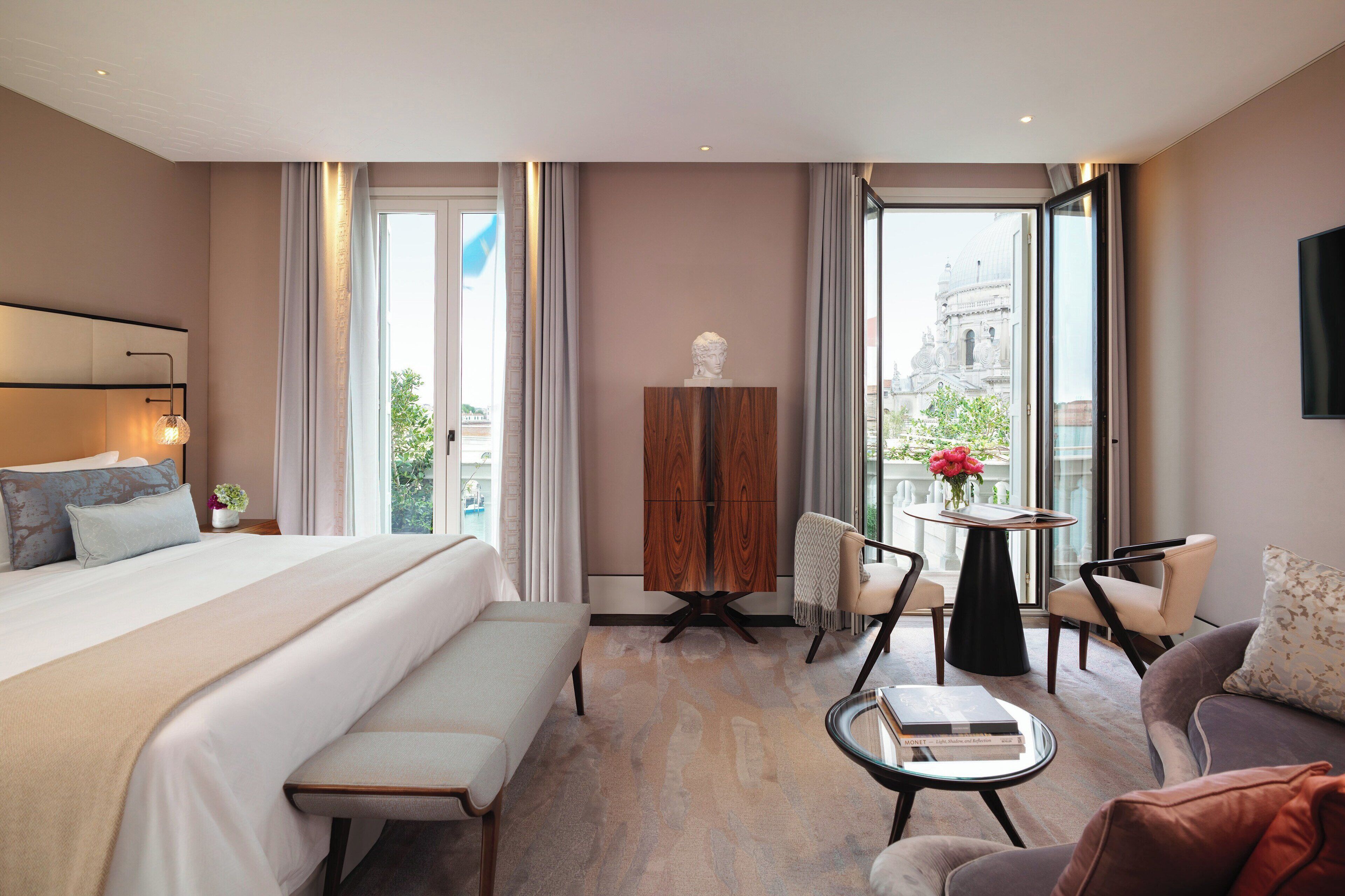 A modern hotel room with a large bed, a sitting area with a bench and chairs, a small table, and a view of a historic building through the window.