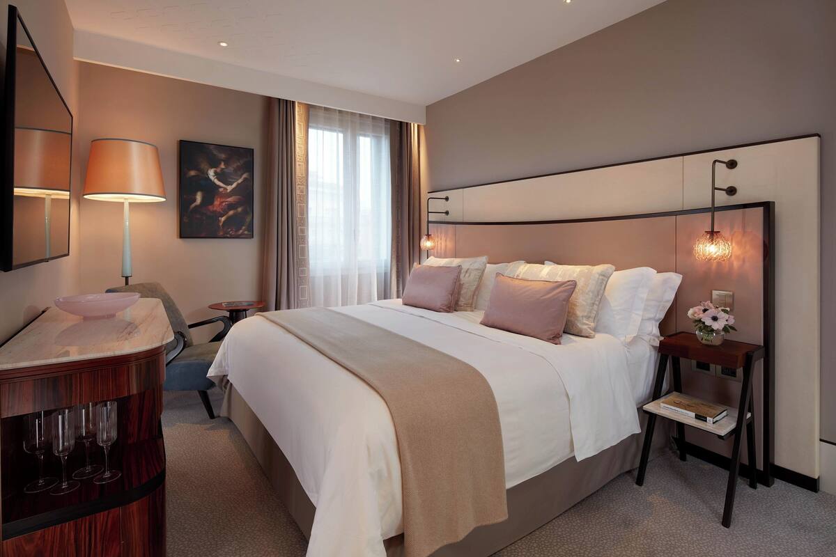 A modern hotel room with a large bed, bedside tables, a desk with a mirror, and a window with curtains.