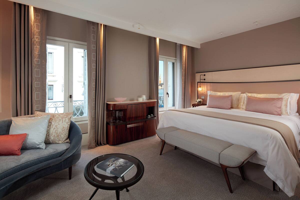 A hotel room with a large bed, a sofa, a bench, a small table, and a wooden cabinet.