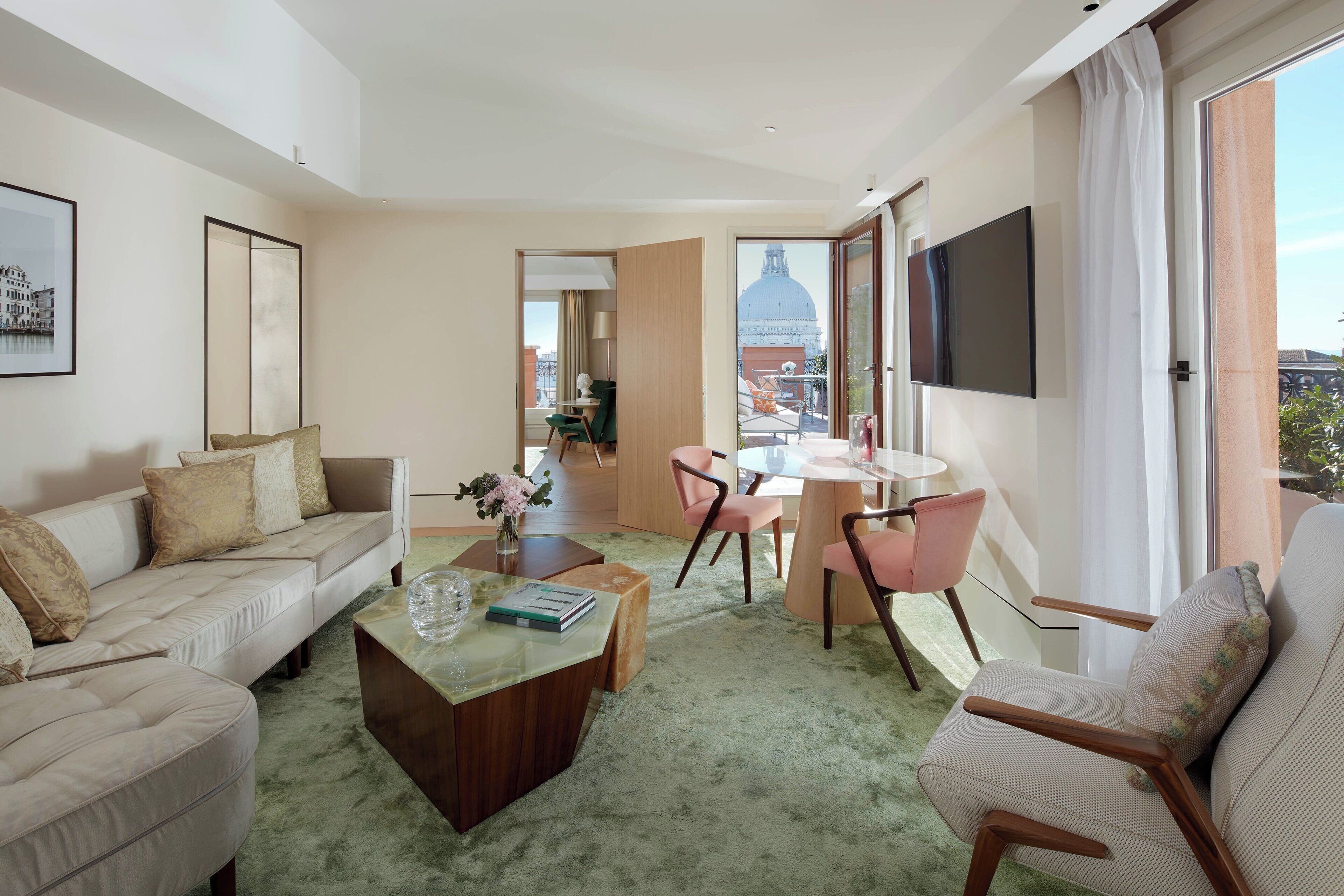 Una sala moderna con sofá, mesa de centro y sillas, un área de comedor con mesa y sillas, y un ventanal con vistas a un edificio.