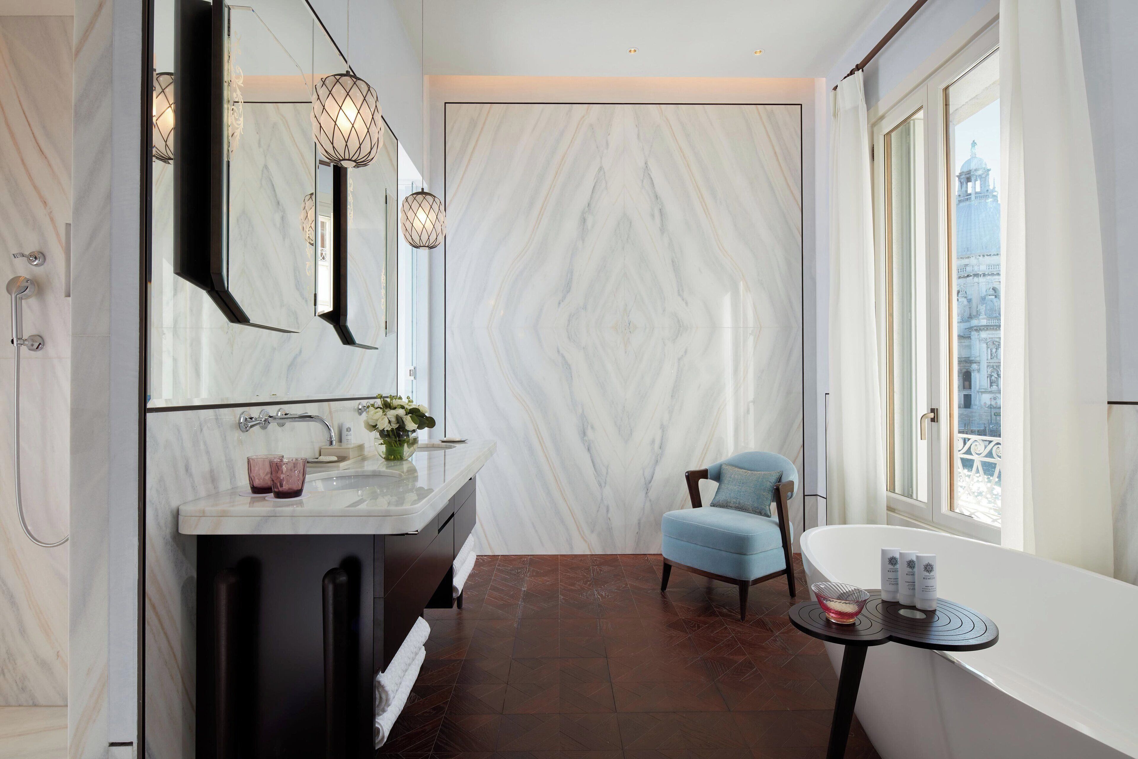 A modern bathroom with a marble wall, a large mirror, a bathtub, and a view of a cityscape through a window.