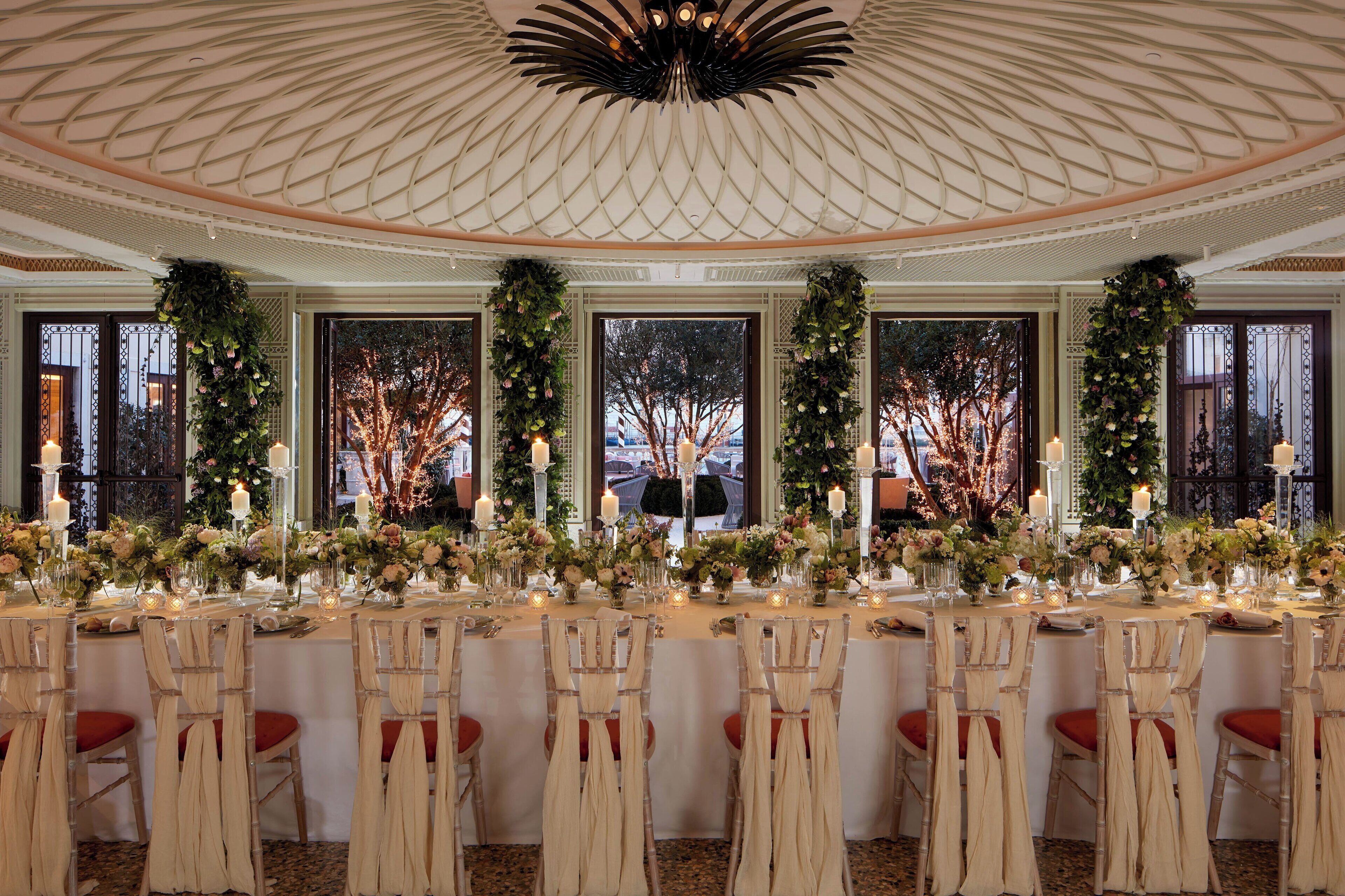 Une salle de banquet dotée d’un plafond haut et de grandes fenêtres, décorée de compositions florales et de bougies.