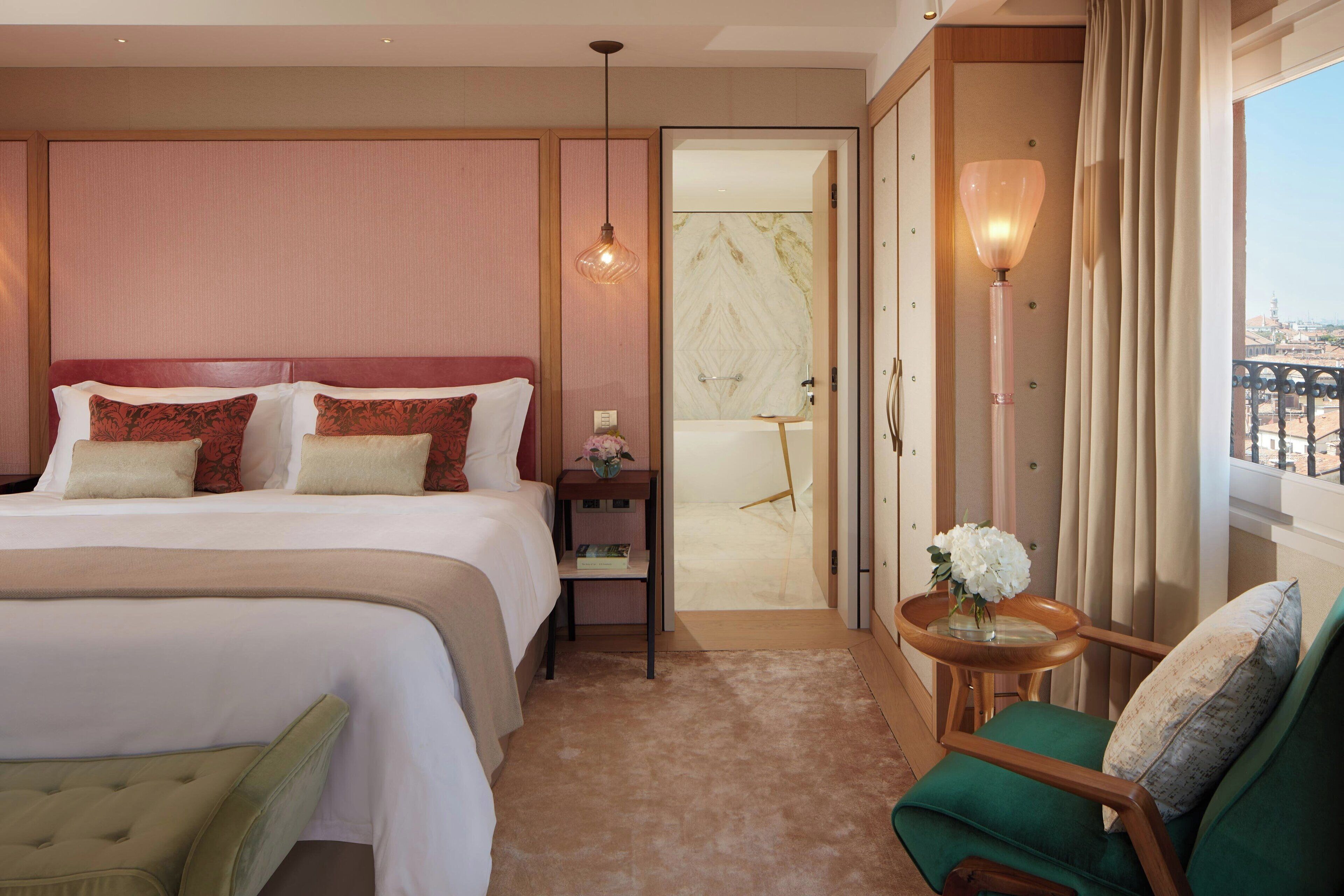 Quarto de hotel com uma cama grande, uma poltrona verde, uma mesinha com um vaso e vista para a cidade pela janela.