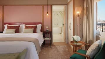 Quarto de hotel com uma cama grande, uma poltrona verde, uma mesinha com um vaso e vista para a cidade pela janela.