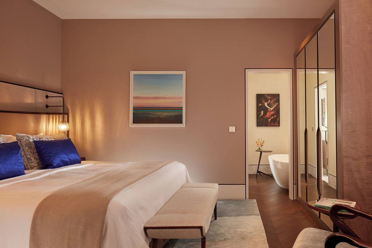 A modern hotel room with a large bed, a bench, a desk, and a view of the bathroom.