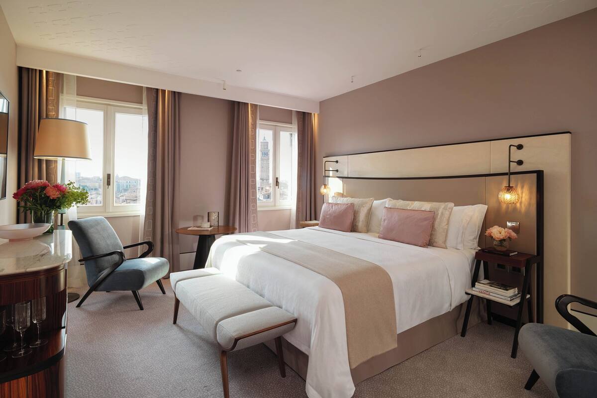 A hotel room with a large bed, a desk with a chair, a bench, and a view of a cityscape through the window.