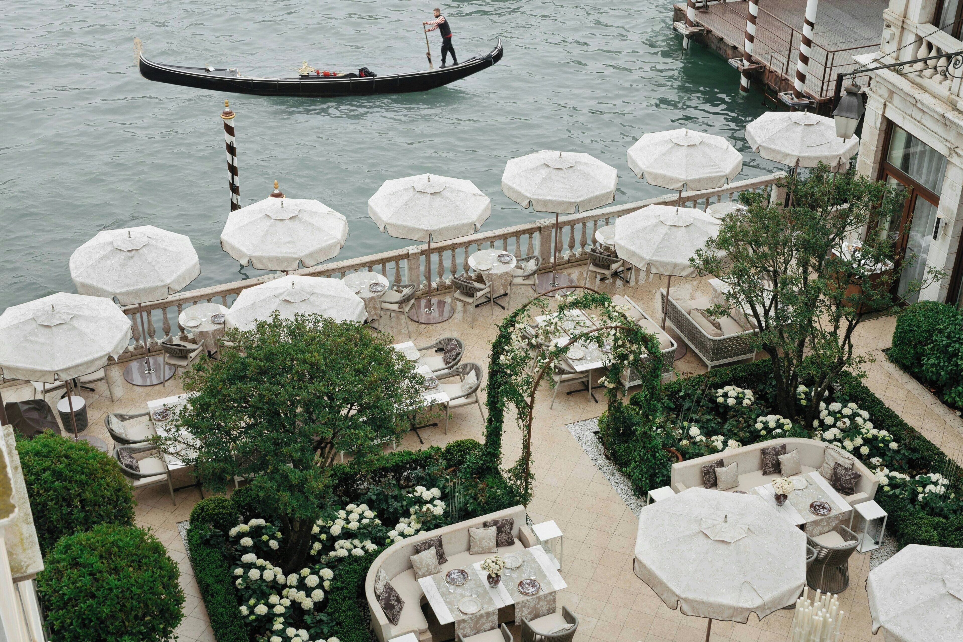 Une terrasse extérieure dotée de plusieurs tables et de parasols, donnant sur un canal où l’on peut apercevoir une gondole.
