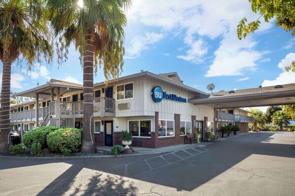 A hotel with a sign reading "BW Best Western." It has a covered outdoor area with a balcony, and there are palm trees and shrubs around.