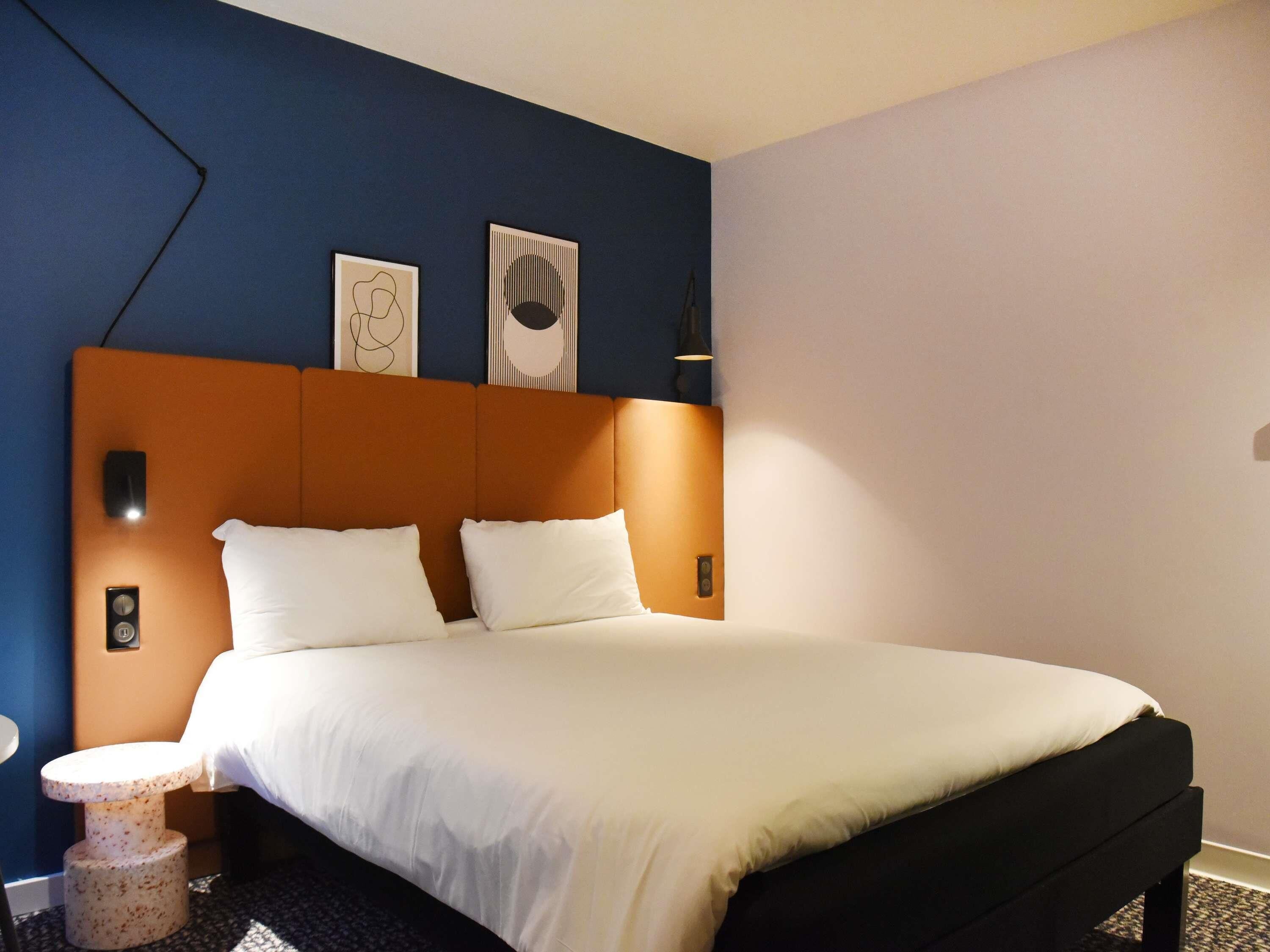A hotel room with a bed, a nightstand, a wall-mounted lamp, and two framed artworks.