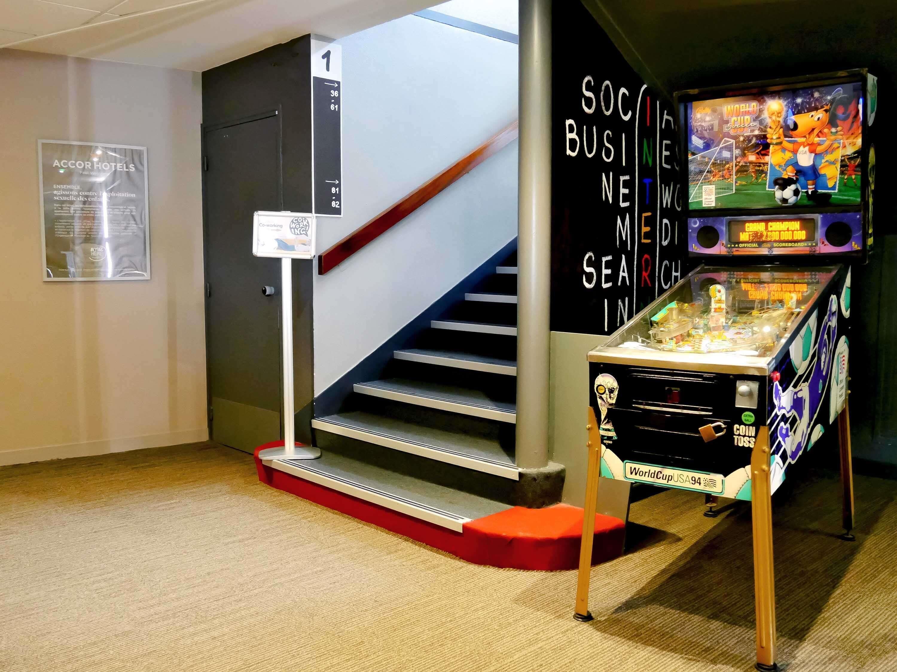 A staircase with a sign indicating the direction to the 1st floor, a coin toss game machine, and a poster for ACCOR Hotels.
