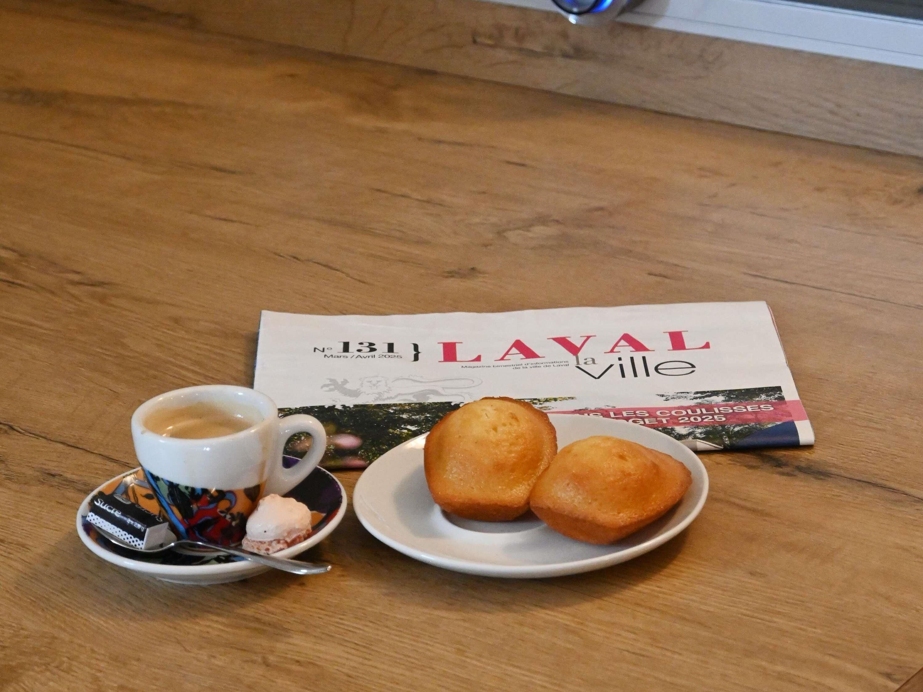 Ein Tisch mit einer Tasse Kaffee, einem Tablett mit zwei Gebäckstücken und einer Zeitung mit der Überschrift 'LAVAL ville'.