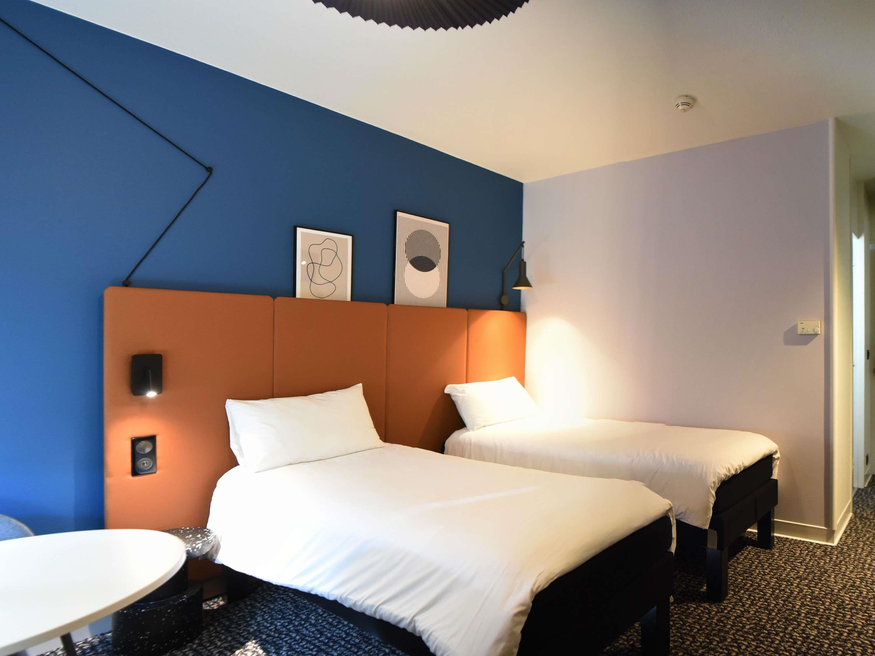 Hotellihuone, jossa on kaksi sänkyä, pieni pyöreä pöytä ja seinään kiinnitetty valaisin.