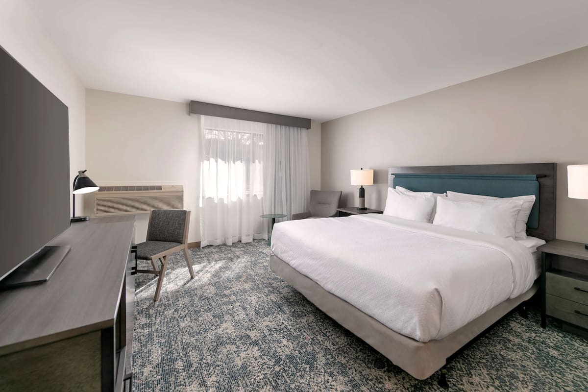 A hotel room with a bed, two bedside tables with lamps, a desk with a chair, a window with curtains, and a wall-mounted air conditioner.