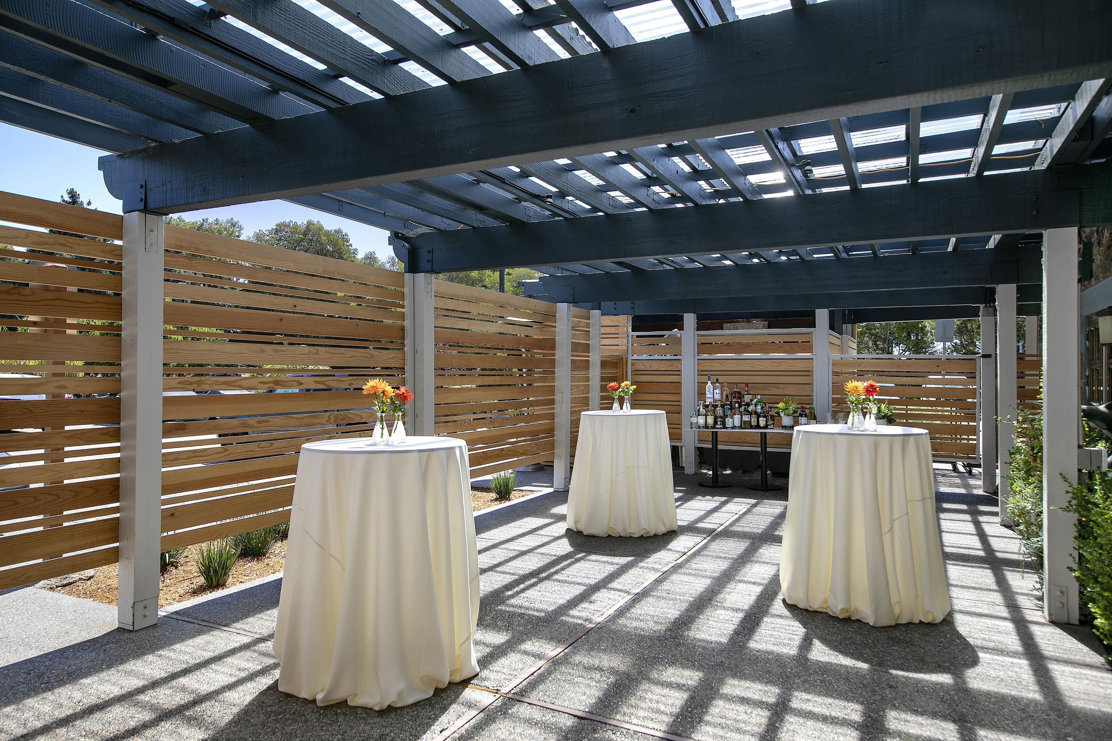 An outdoor patio area with a pergola structure, wooden slats, and round tables with white tablecloths.