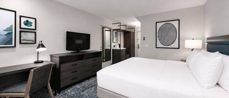 Hotellihuone, jossa on sänky, työpöytä tietokoneella, tuoli, televisio ja taideteoksia seinillä.