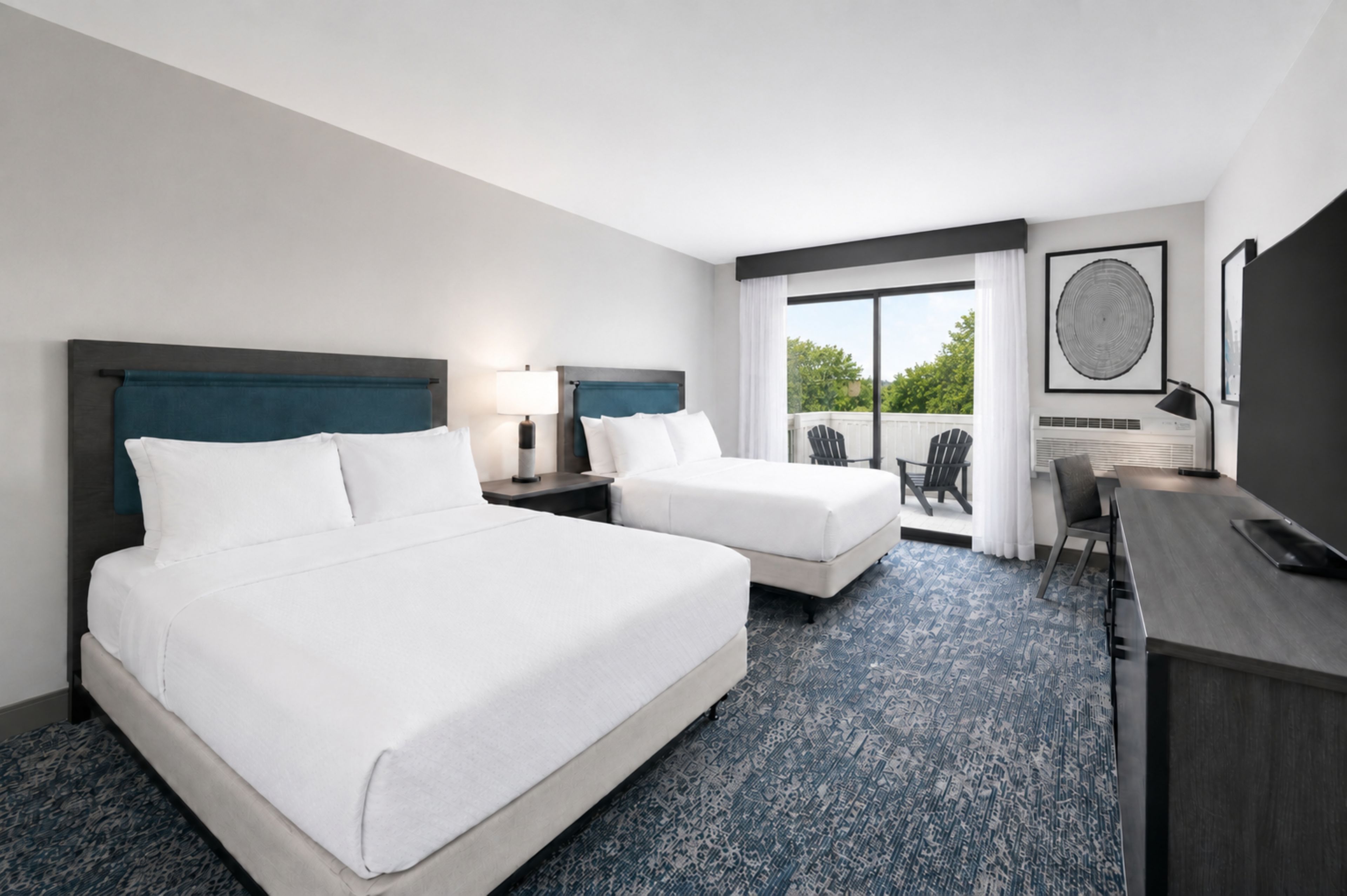 A hotel room with two beds, a desk, a chair, and a balcony with a view of trees.
