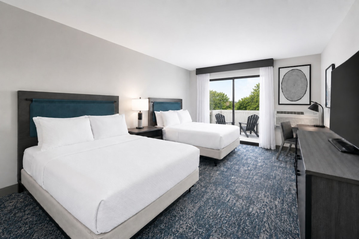 A hotel room with two beds, a desk, a chair, and a balcony with a view of trees.