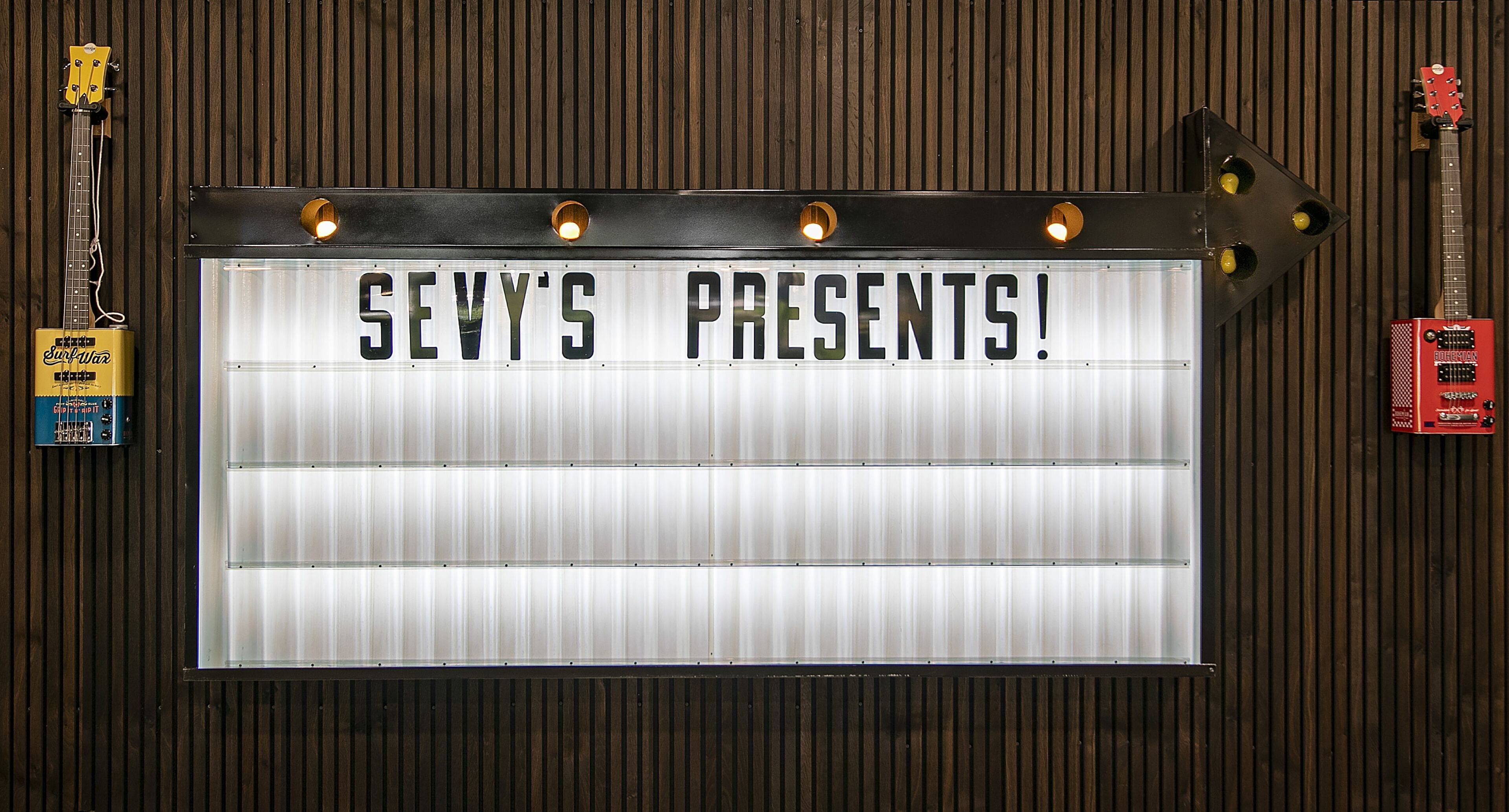 Une enseigne portant l’inscription « SEVY’S PRESENTS ! » est fixée à un mur, et de chaque côté de l’enseigne sont accrochées deux guitares.