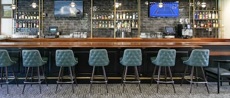 Zona de bar con una barra larga, varios taburetes y una pantalla de televisión mostrando un partido de tenis.