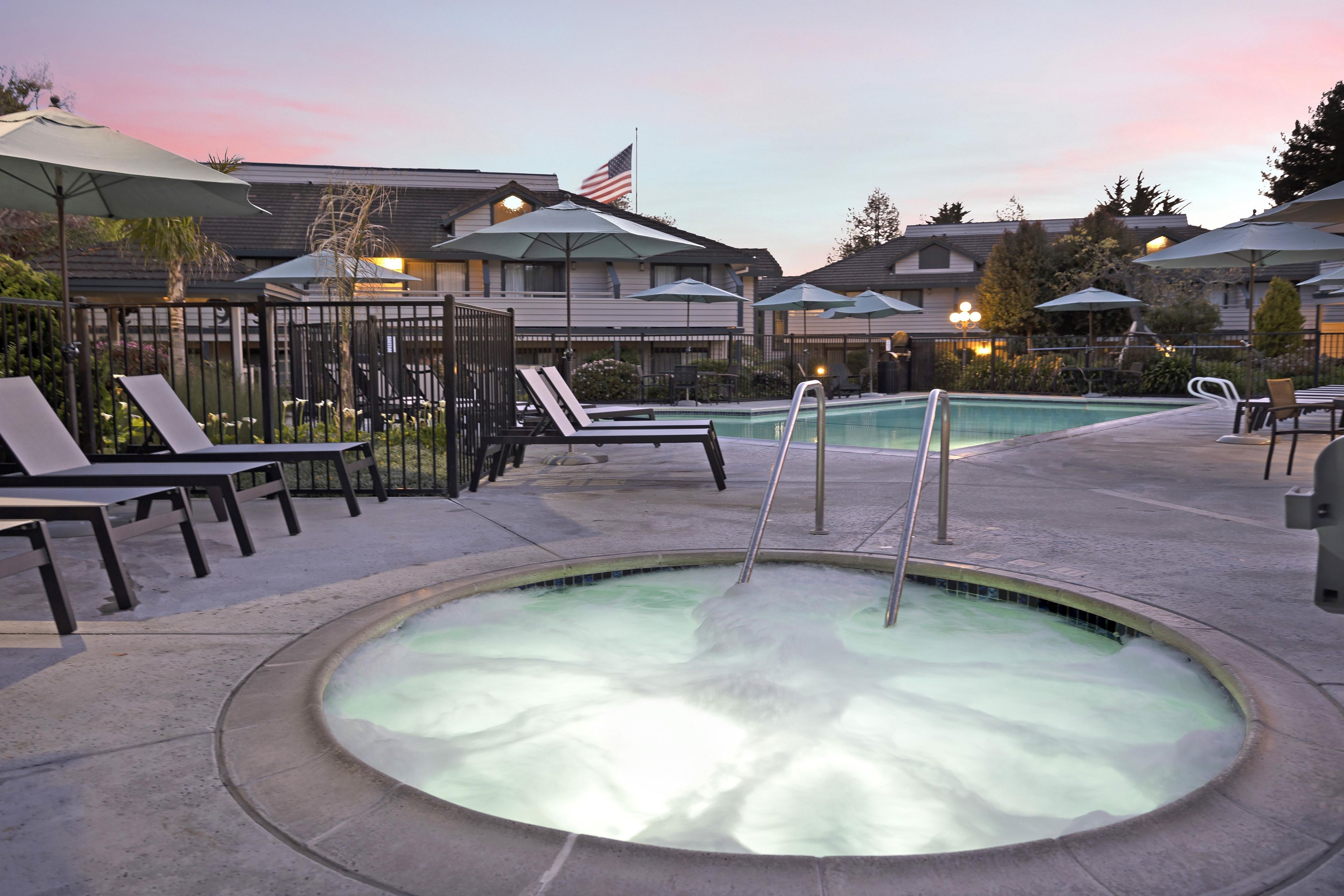 A hot tub surrounded by lounge chairs and umbrellas, with a pool in the background and a flag on a building.