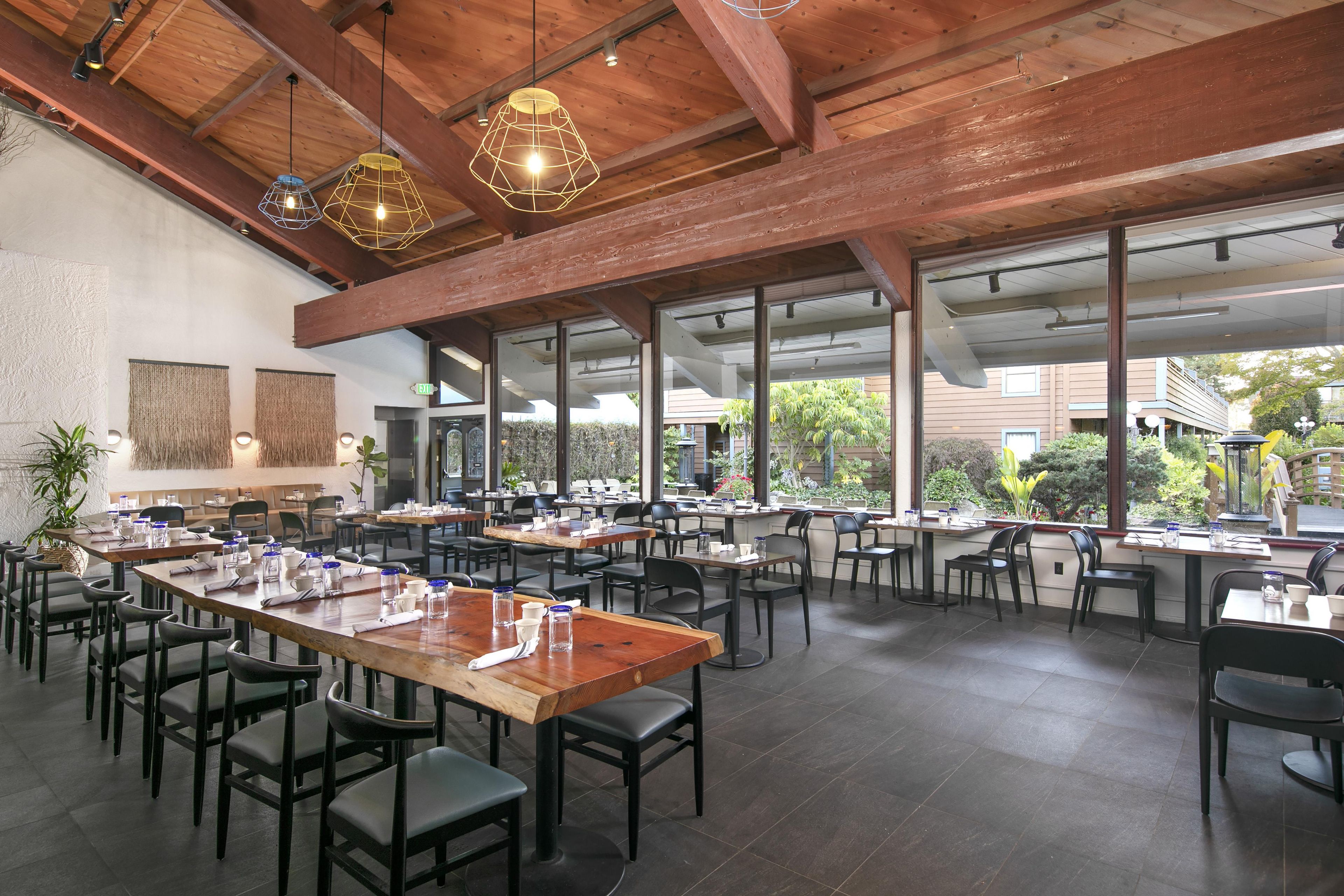 A spacious dining area with multiple tables and chairs, large windows offering a view of the outdoors, and a high ceiling with exposed wooden beams.