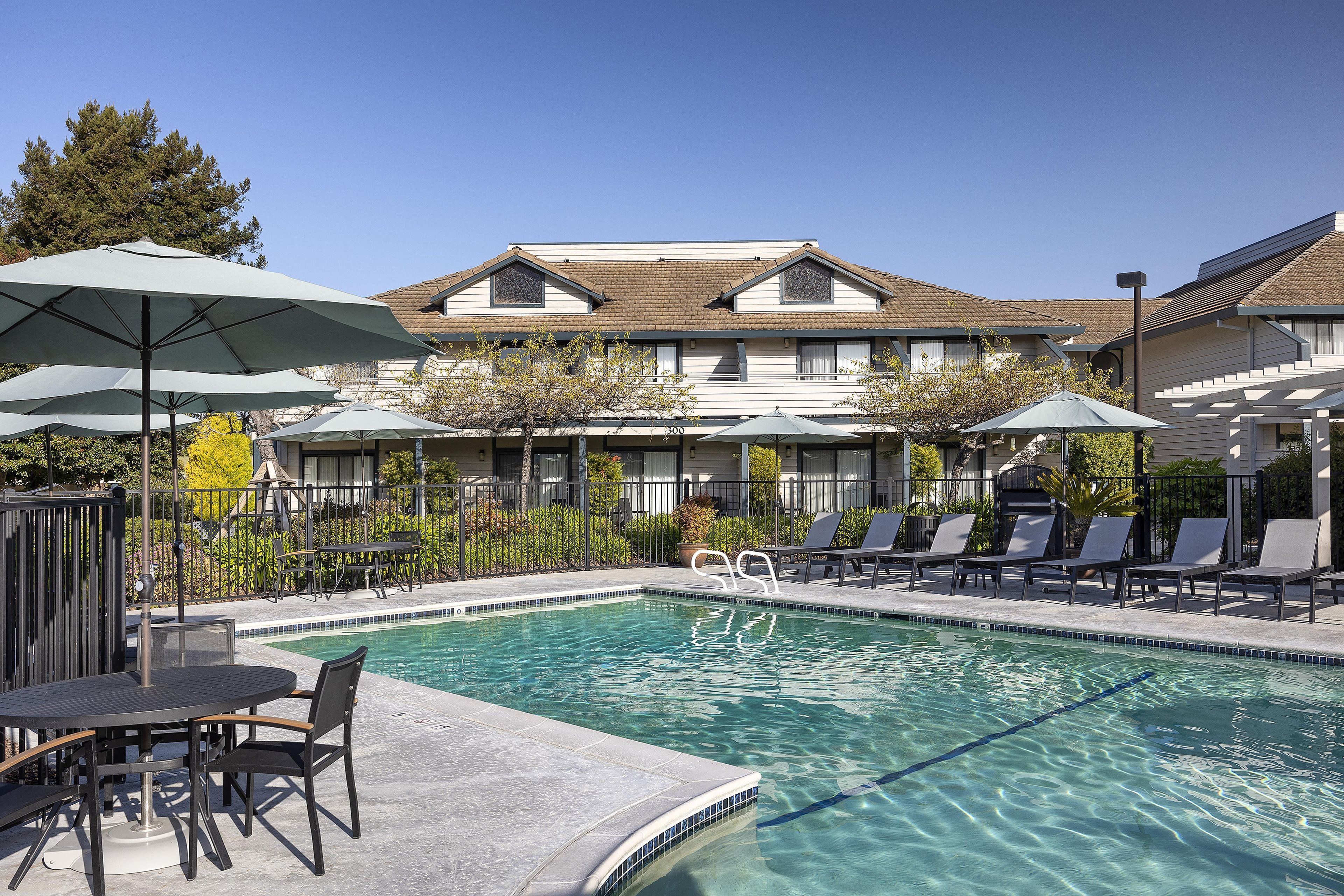 A swimming pool with lounge chairs and umbrellas, surrounded by a fence and trees.