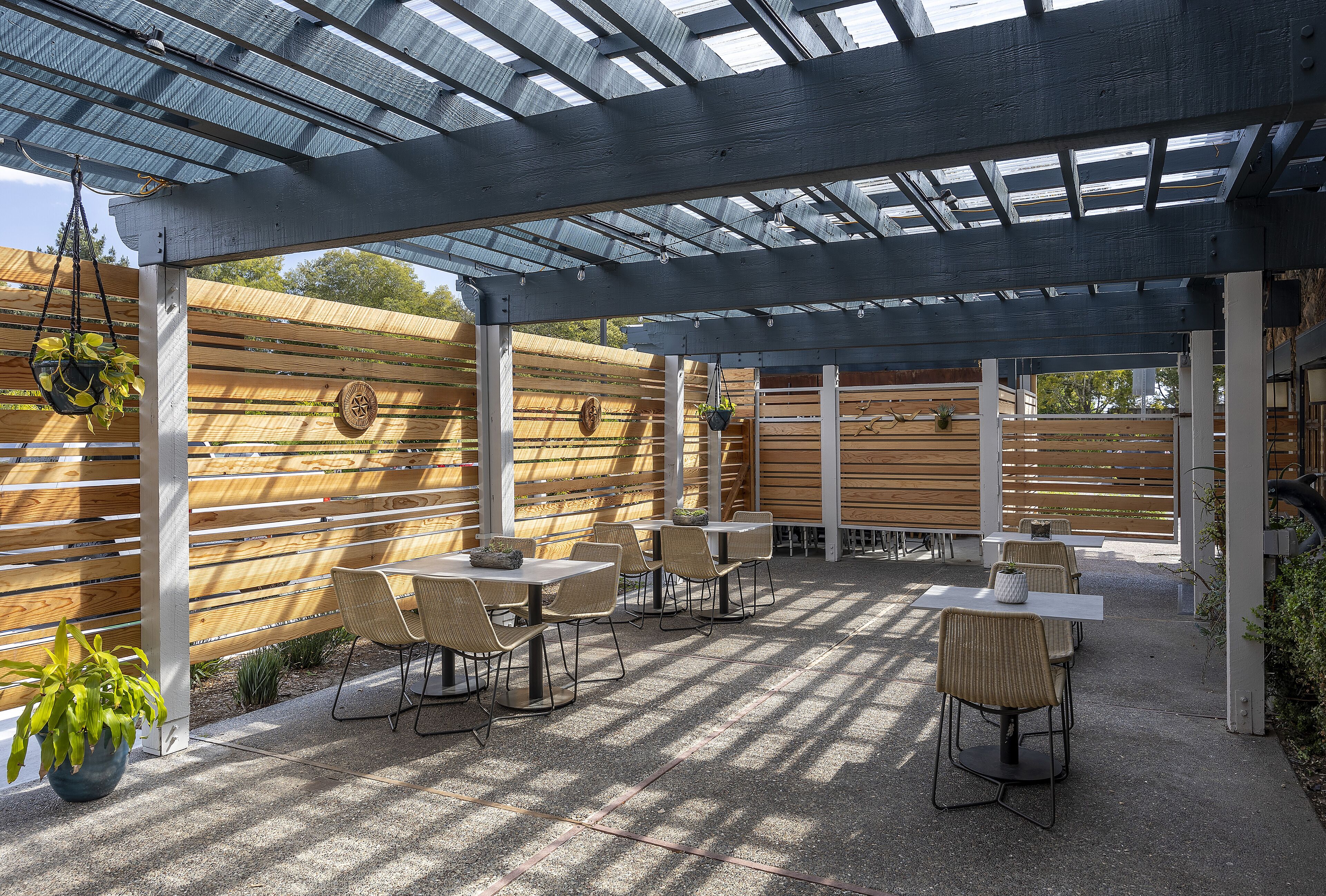 An outdoor patio with tables and chairs, a wooden fence, and a pergola with a glass roof.