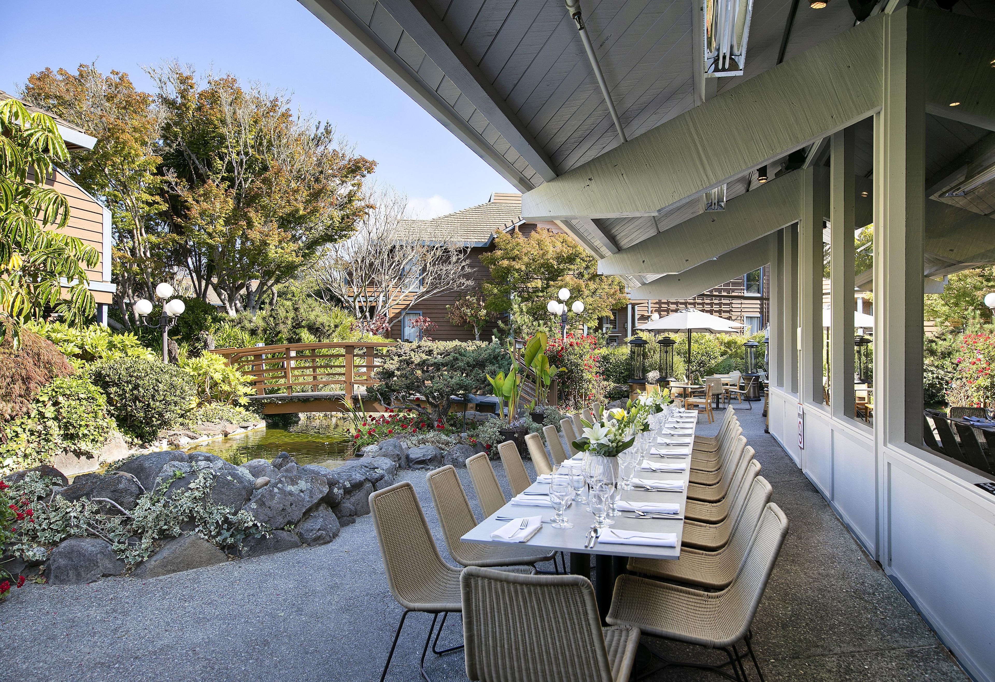 Un área de comedor al aire libre con mesas y sillas, rodeada por un jardín con rocas y plantas.