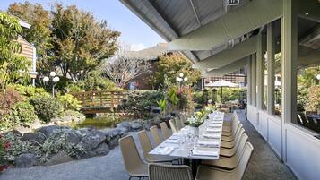 Un área de comedor al aire libre con mesas y sillas, rodeada por un jardín con rocas y plantas.
