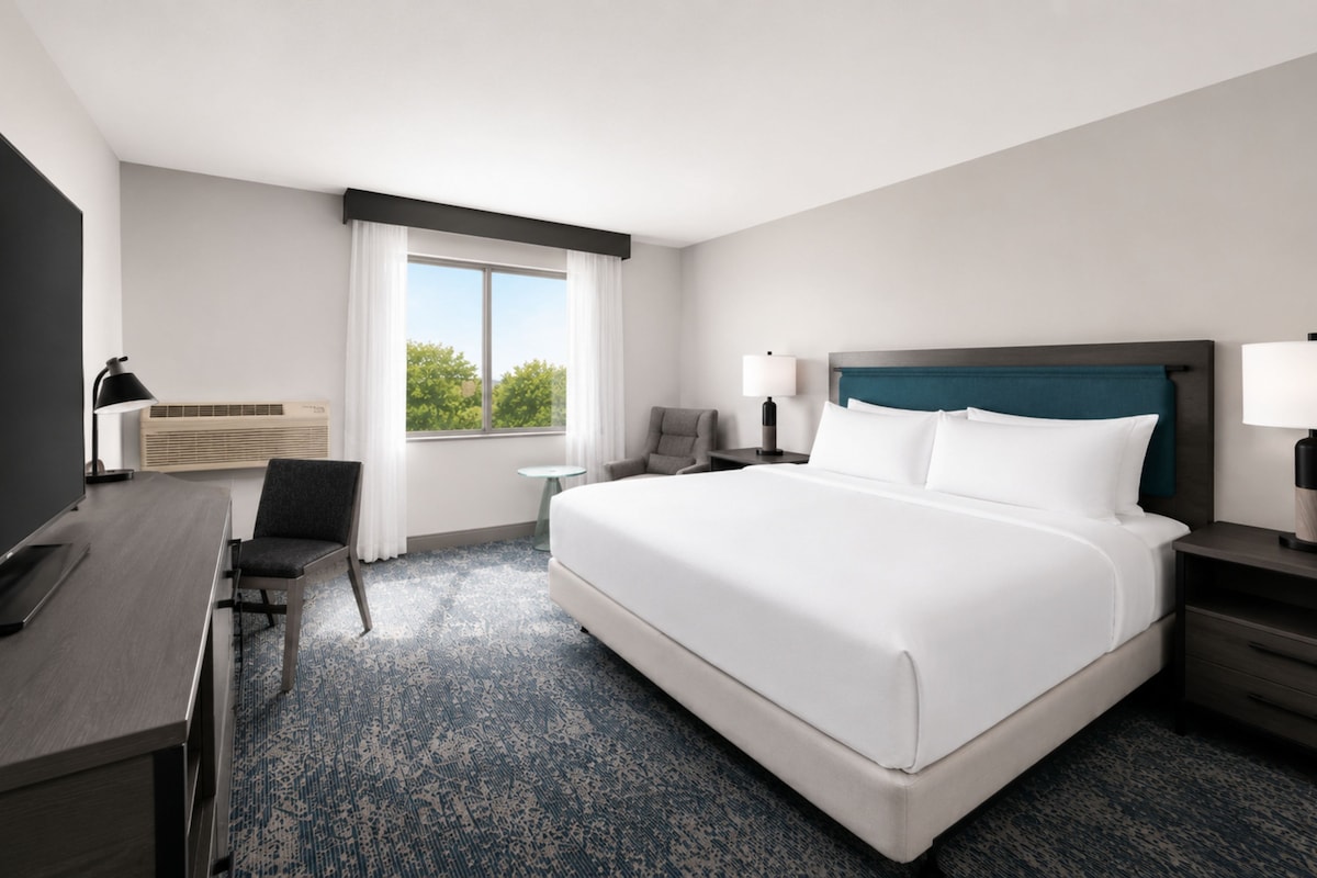 A hotel room with a large bed, a desk, a chair, a nightstand, a lamp, and a window with a view.
