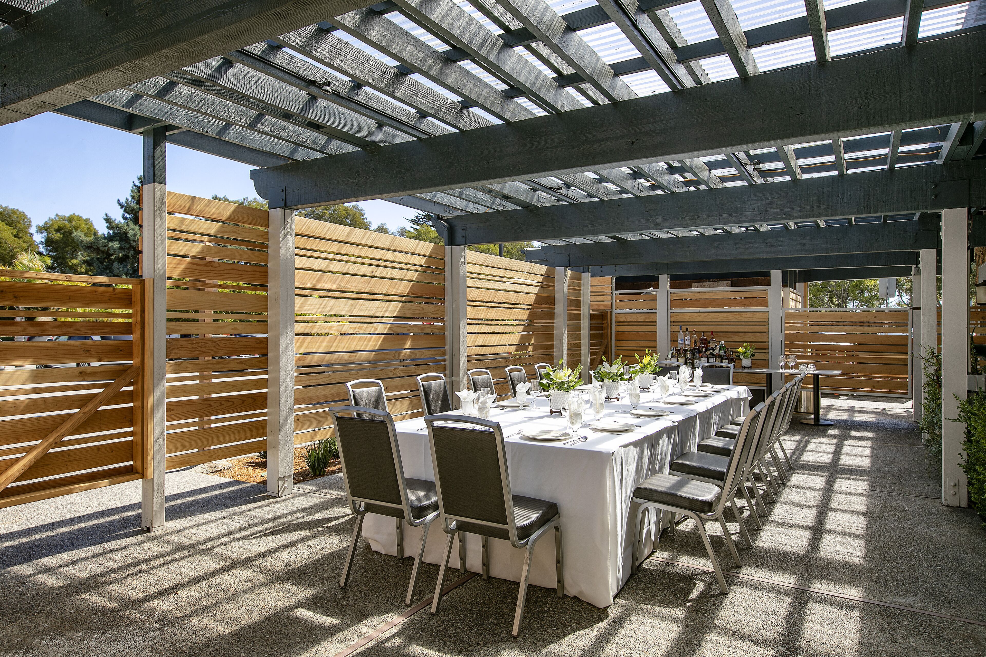 Un patio al aire libre con una larga mesa preparada para comer, rodeada de sillas, bajo una estructura de pérgola.