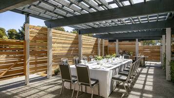 Un patio al aire libre con una larga mesa preparada para comer, rodeada de sillas, bajo una estructura de pérgola.