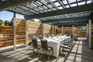Un patio al aire libre con una larga mesa preparada para comer, rodeada de sillas, bajo una estructura de pérgola.