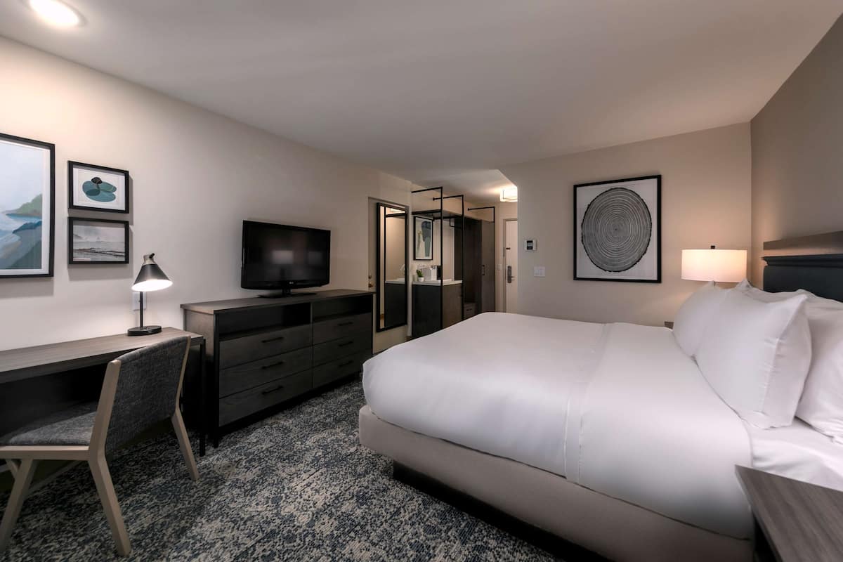 A hotel room with a bed, desk, chair, TV, dresser, and artwork on the walls.