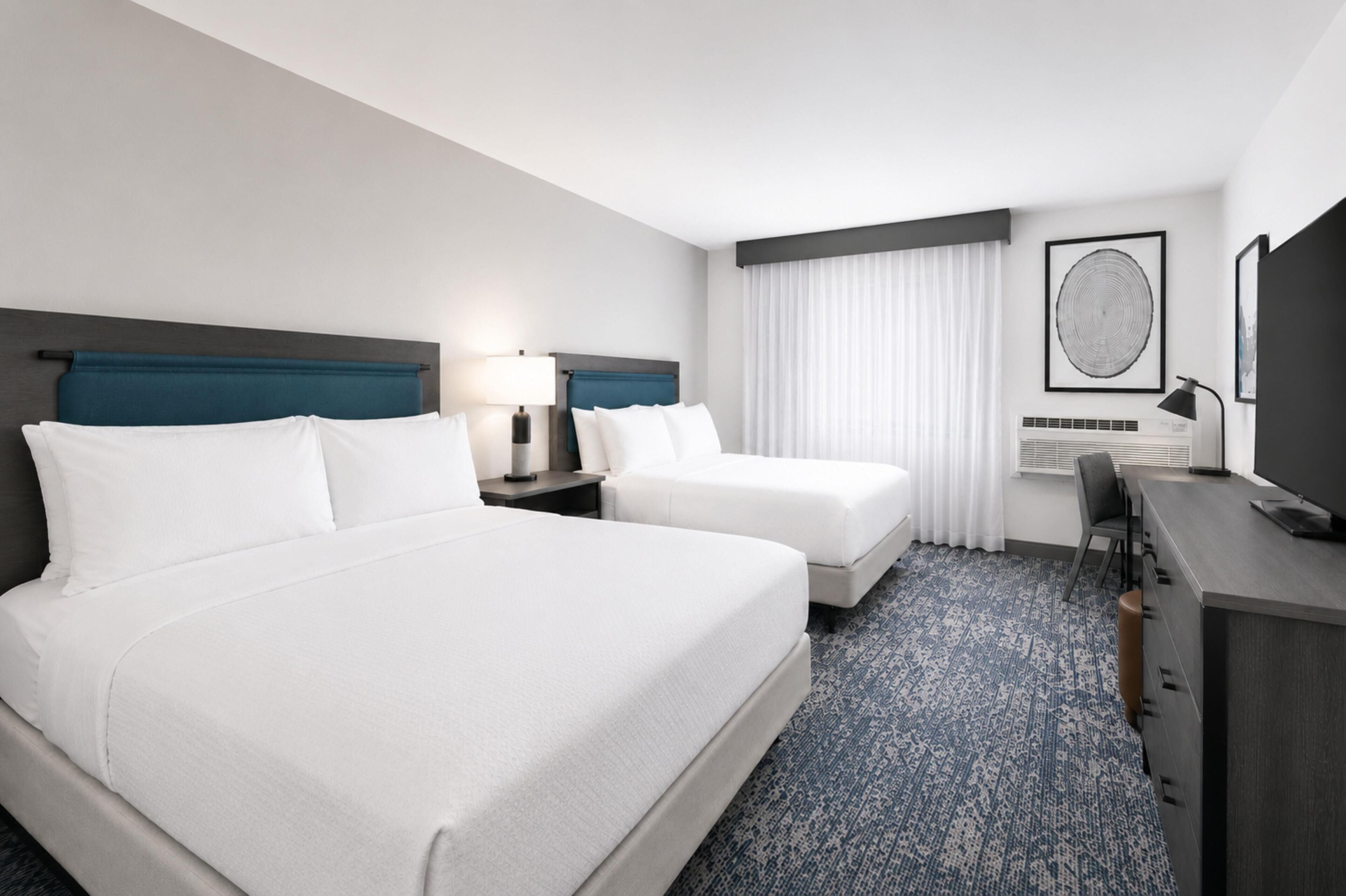 Quarto de hotel com duas camas, uma escrivaninha, uma cadeira, um abajur e uma janela grande com cortinas.